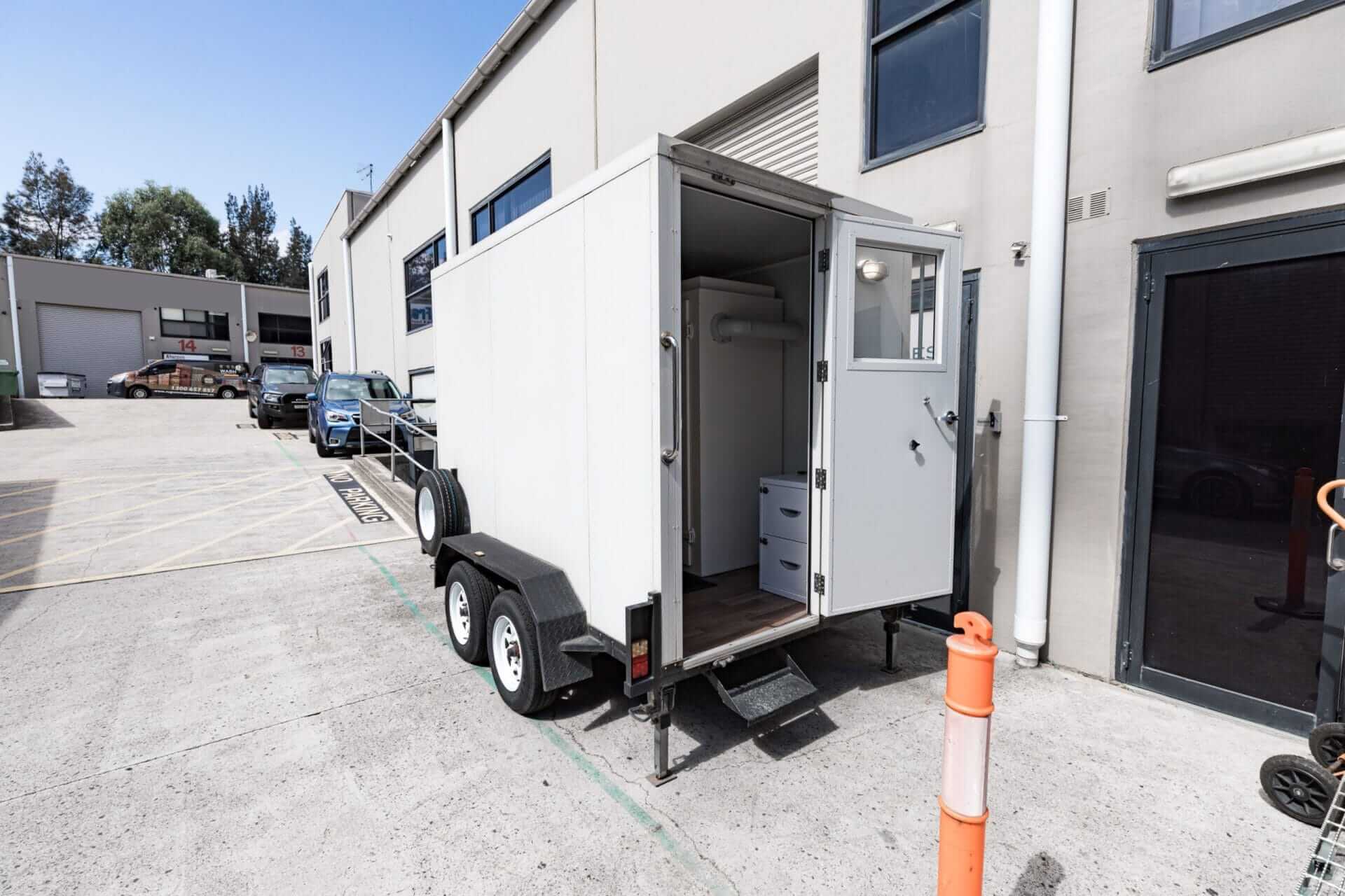 A portable toilet trailer is parked in front of a workplace building.